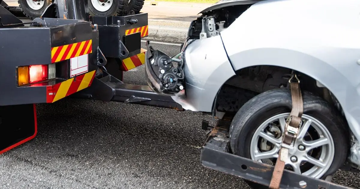 A damaged silver car is secured on a tow truck after a crash, with visible front-end impact.