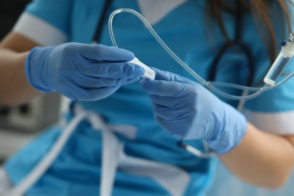 Nurse wearing blue scrubs and gloves preparing an intravenous catheter for a medical procedure.