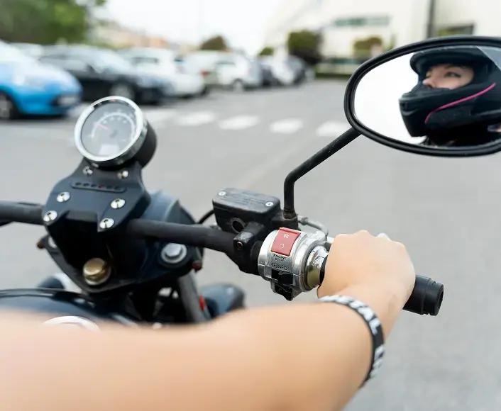 Motorcyclist wearing a helmet reflected in the rearview mirror, holding the handlebars and preparing to ride through a parking area.