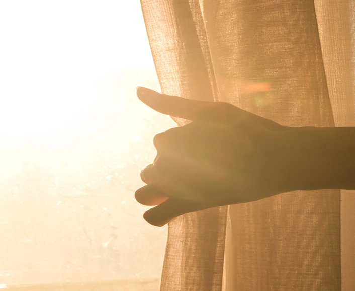 Hand gently touching sunlight through curtain, symbolizing healing and hope after trauma.