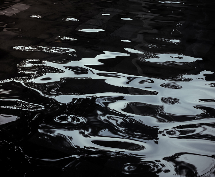 Dark water surface reflecting light, showing the slick, distorted patterns of an oil spill contaminating the environment.