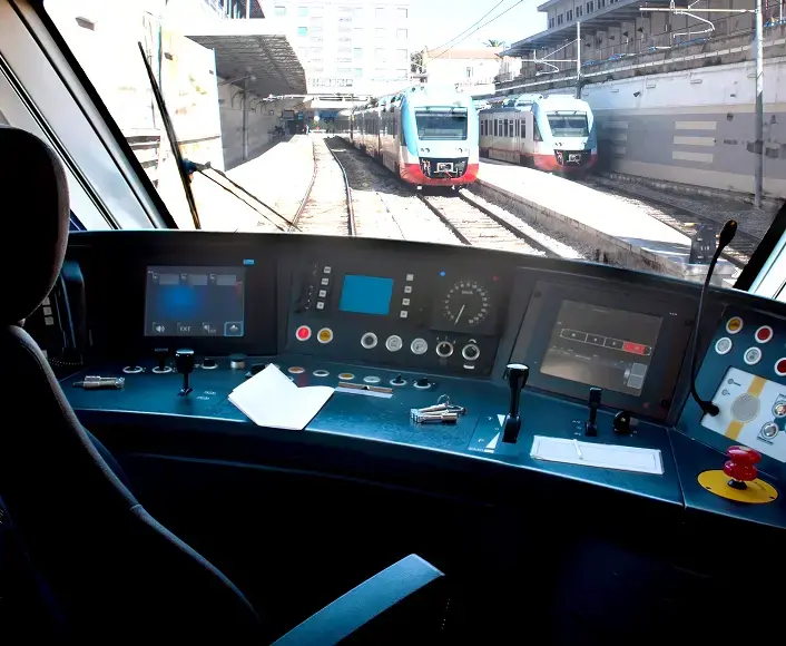 View from inside a modern train cockpit looking out toward train tracks and two trains at a station, symbolizing transportation and safety oversight.